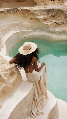 a woman in a white dress and hat sitting on a ledge next to a pool