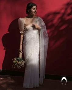 a woman wearing a white sari and holding a bouquet in front of a red wall