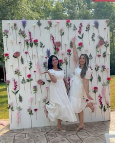 two women in white dresses standing next to a wall with pink flowers and greenery on it