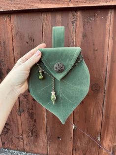 a person holding up a green leaf shaped pouch with charms attached to it, on top of a wooden fence