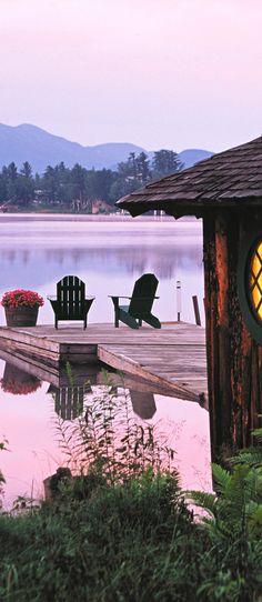 there is a clock on the side of a dock next to some chairs and flowers