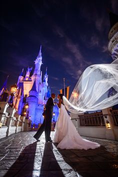 a bride and groom standing in front of a castle at night with their veil blowing in the wind