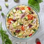 a glass bowl filled with pasta salad next to cucumbers, tomatoes and herbs