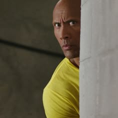 a bald man in a yellow shirt looking out from behind a white wall with his hands on his hips