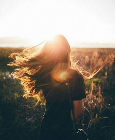 a woman walking through a field with her hair blowing in the wind and sun shining behind her