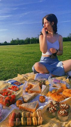 a woman sitting on a blanket in the middle of a field with many different types of food