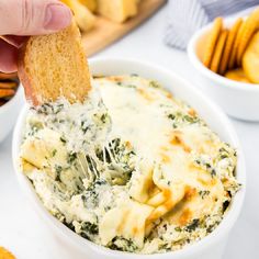 a hand holding a piece of bread over a bowl of spinach artichoke dip