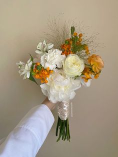 a bouquet of white and orange flowers being held by a person's hand in front of a wall