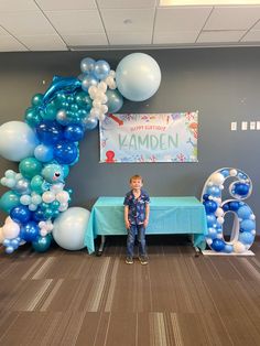 a young boy standing in front of a table with blue and white balloons on it