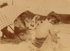 two women sitting on the beach with an umbrella over their heads and one holding a dog