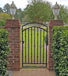 an iron gate in the middle of a brick walkway leading to a grassy area with trees and bushes