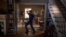 a man standing in the middle of a living room next to a stair case and bookshelf