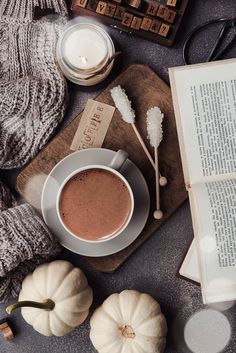 a cup of hot chocolate next to an open book and some pumpkins on a table