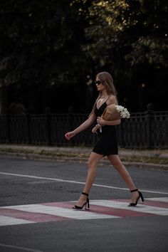 a woman walking across a cross walk holding a flower in her right hand and wearing high heels