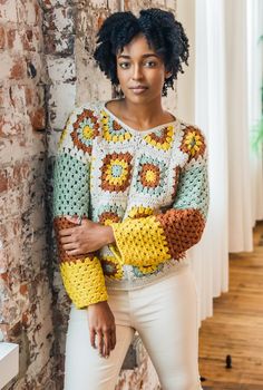 a woman standing in front of a brick wall wearing a colorful crochet sweater