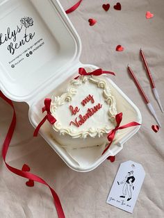 a heart shaped cake in a white box with red ribbon on the table next to it