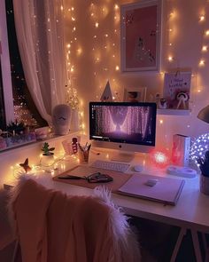 a white desk topped with a computer monitor next to a window covered in fairy lights