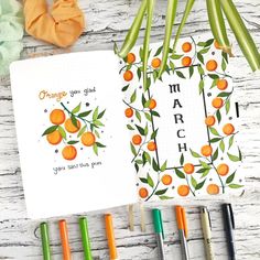 an open notebook with oranges on it next to some markers and pens in front of them