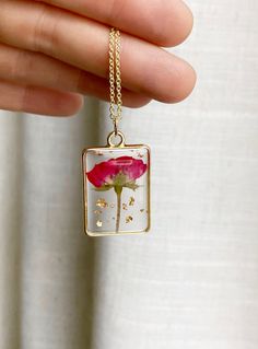 a hand holding a square necklace with a flower in the center and gold flecks on it