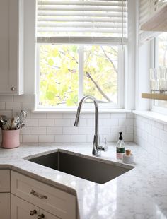 a white kitchen with marble counter tops and sink under a window that has blinds in it