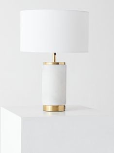 a white table lamp with a gold base on top of a white shelf next to a white wall