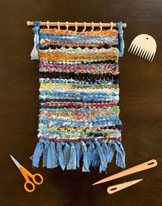 a weaving project with scissors, combs and yarn on a wooden table next to it