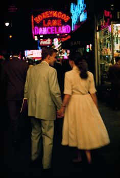 a man and woman holding hands walking down the street in front of a neon sign