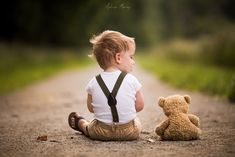 a little boy sitting in the middle of a road with a teddy bear next to him