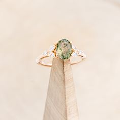 an engagement ring with a green stone surrounded by white diamonds on top of a wooden stand