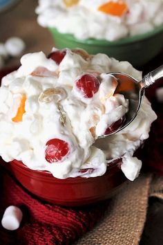 two bowls filled with whipped cream and cherries on top of a red cloth next to a spoon