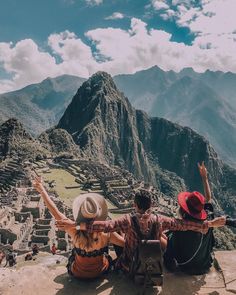 two people sitting on the edge of a cliff with their arms in the air and mountains in the background