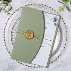 a green wedding card with a gold button on it sitting on a plate next to flowers