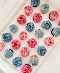 cupcakes with pink, blue and gold frosting in a white box on a table