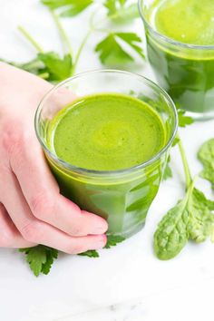 two glasses filled with green smoothie on top of a white table next to leaves