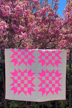 a pink and gray quilt hanging on a fence next to trees with blossoming flowers