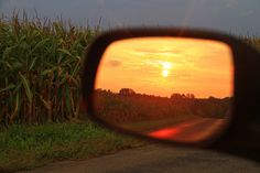 the sun is setting in front of a car's rear view mirror as it travels down a country road