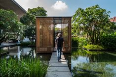 a man walking across a bridge over a pond next to a wooden structure with bamboo covering it