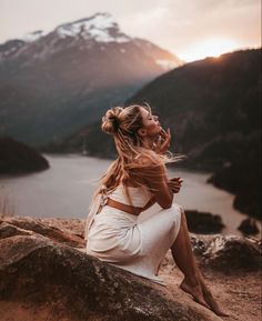 a woman sitting on top of a large rock next to a body of water with mountains in the background