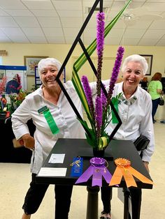 two women standing next to each other with flowers in front of them and ribbons on the table