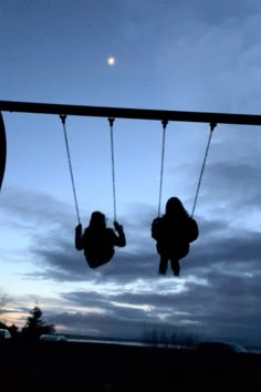 two people sitting on swings in front of the sky at dusk, with one person hanging upside down