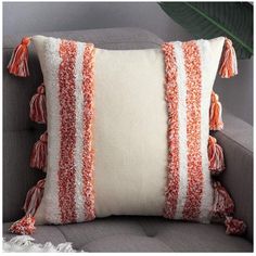 an orange and white pillow with tassels sits on a gray couch next to a plant