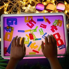 two hands are playing with letters and numbers on a lit up tablet screen in front of christmas lights