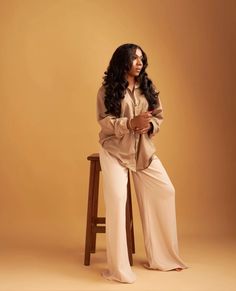 a woman sitting on top of a wooden chair in front of a brown wall wearing long pants