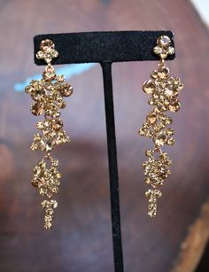 a pair of gold colored earrings sitting on top of a black stand in front of a wooden