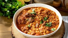 a white bowl filled with meat and bean stew on top of a wooden table next to parsley