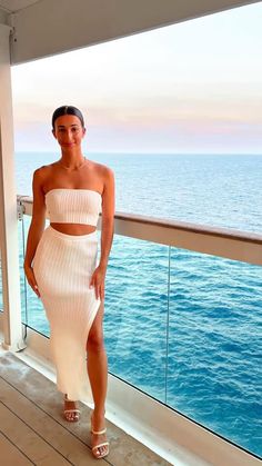 a woman standing on top of a balcony next to the ocean wearing a white dress