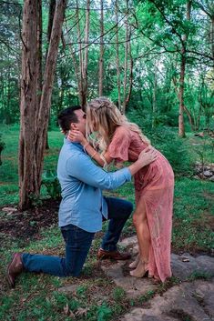 a man kneeling down to kiss a woman's face in the woods with trees behind them