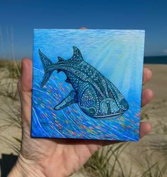 a hand holding up a painting of a shark on the beach with blue sky and clouds in the background