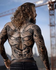 a man with long hair and tattoos standing in front of a construction site looking at the sky