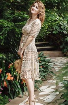 a woman in a dress is standing on a stone walkway with flowers and trees behind her